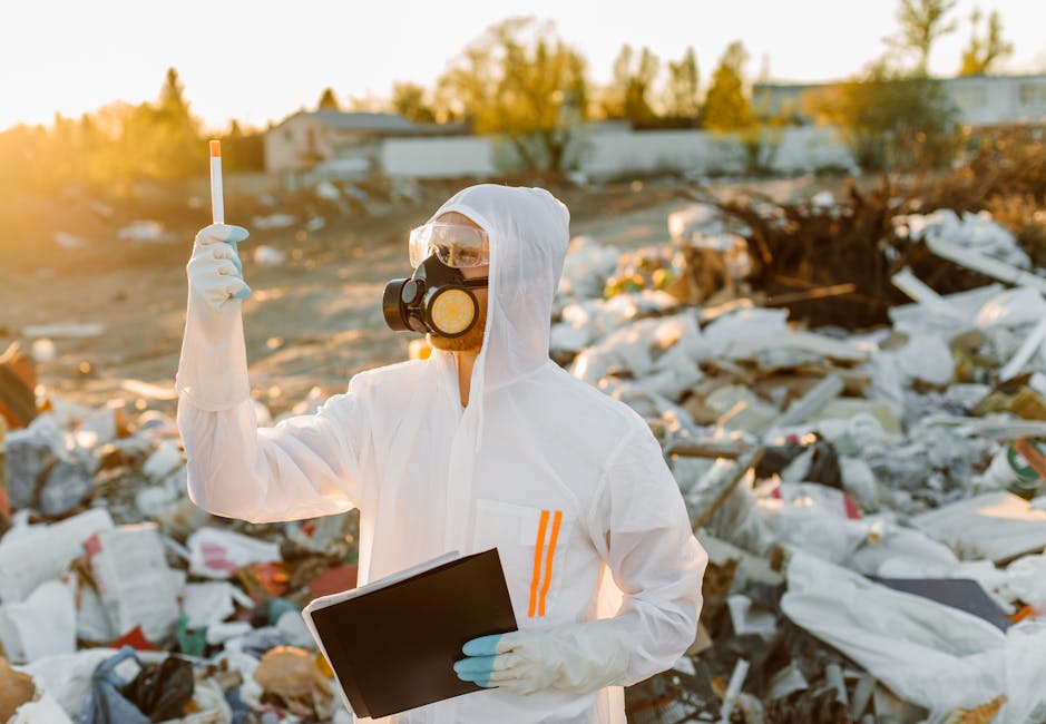 A person dressed in a white protective suit with orange reflective stripes, wearing black gloves, a hood, and goggles, stands amidst a large pile of mixed household waste and rubbish on a beach or lan