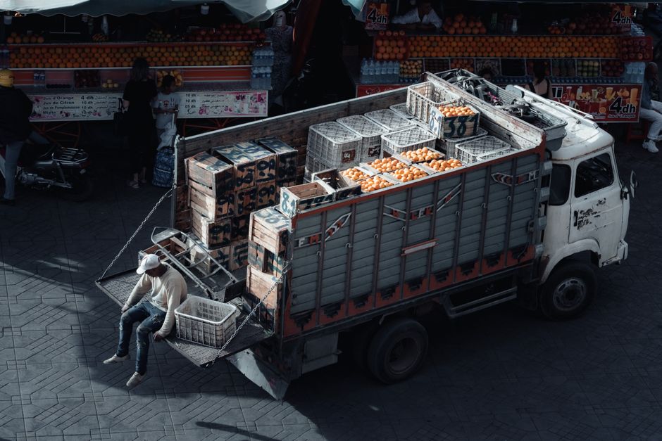 A white delivery truck with an open cargo area is parked on a paved street, loaded with wooden crates and plastic containers filled with oranges and other produce, ready for transport. A man wearing a