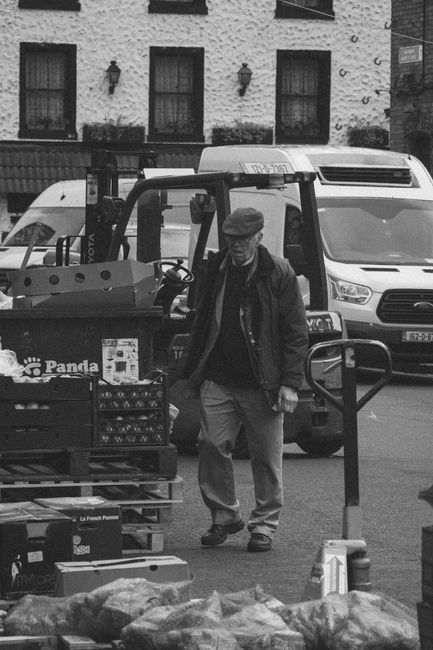 A white delivery truck with an open cargo area is parked on a paved street, loaded with wooden crates and plastic containers filled with oranges and other produce, ready for transport. A man wearing a